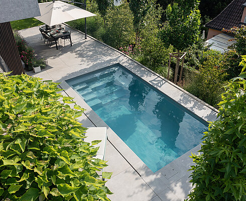 Privatgarten B Moderner rechteckiger Swimmingpool in einem Garten, umgeben von grünen Pflanzen, mit einer Terrasse und einem Tisch mit Sonnenschirm im Hintergrund.